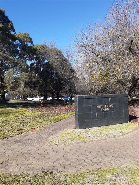 Stone wall of entrance to Settlers park, with path surrounding wall and trees in the background