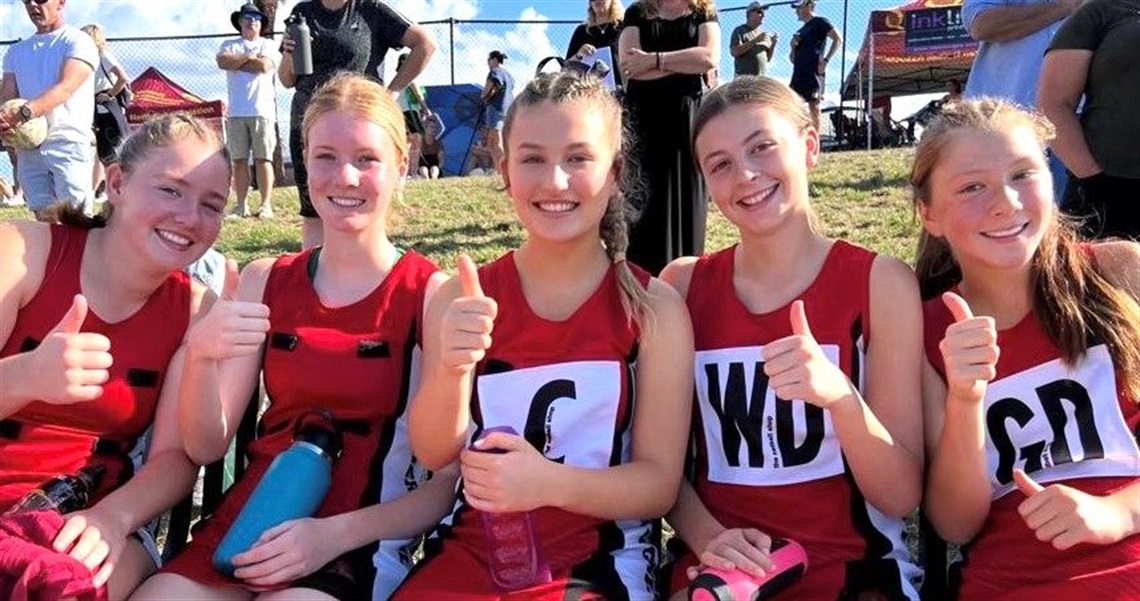 5 young women in netball uniforms sitting court side, smiling and giving thumbs up to camera.