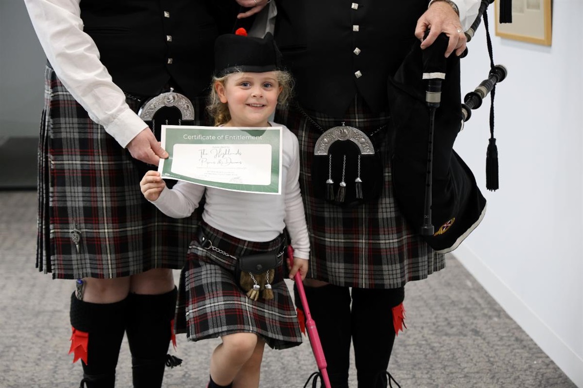 Family of three wearing kilts and young girl holds up certificate