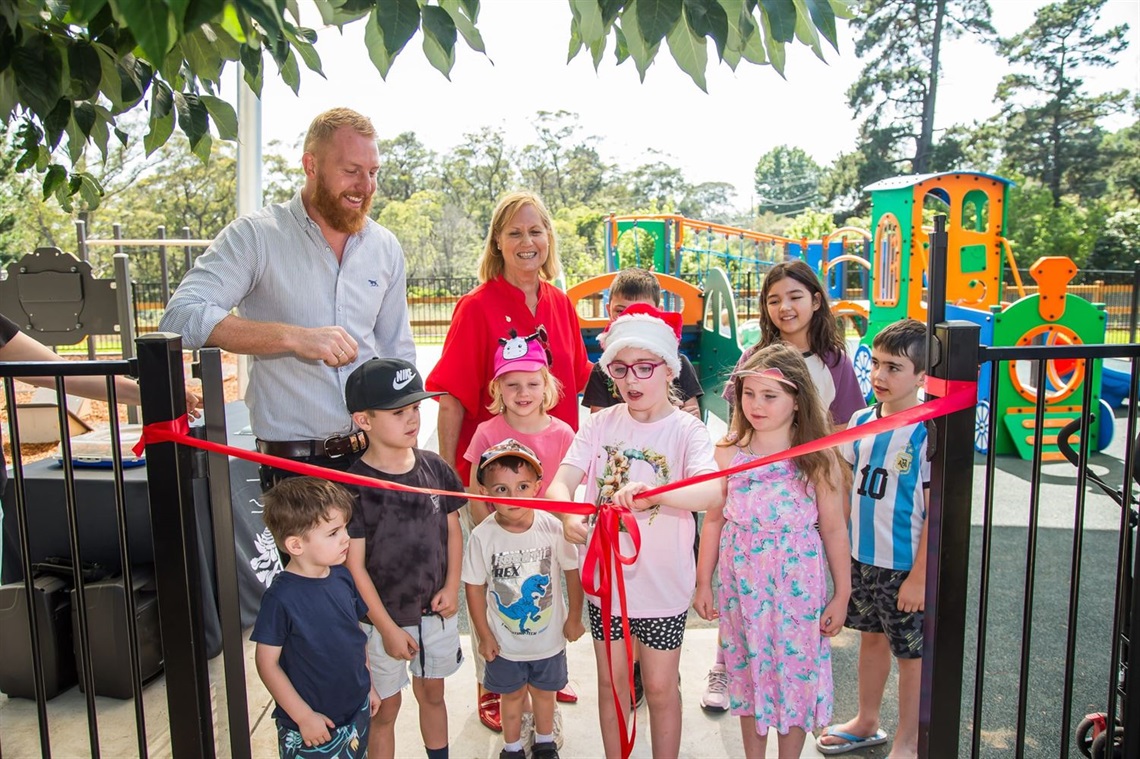 Young girl with Christmas hat cuts red ribbon at the gate entrance of Hill Top Community Park with seven other children as well as Mayor Jesse Fitzpatrick and Judy Hannan MP 