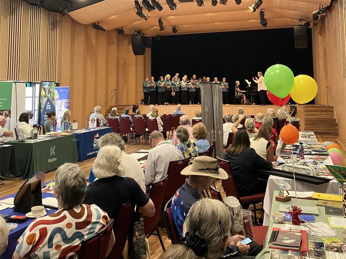 Seniors Festival Expo - Long Tables, guest seating and information tables in hall with Choir performing on stage