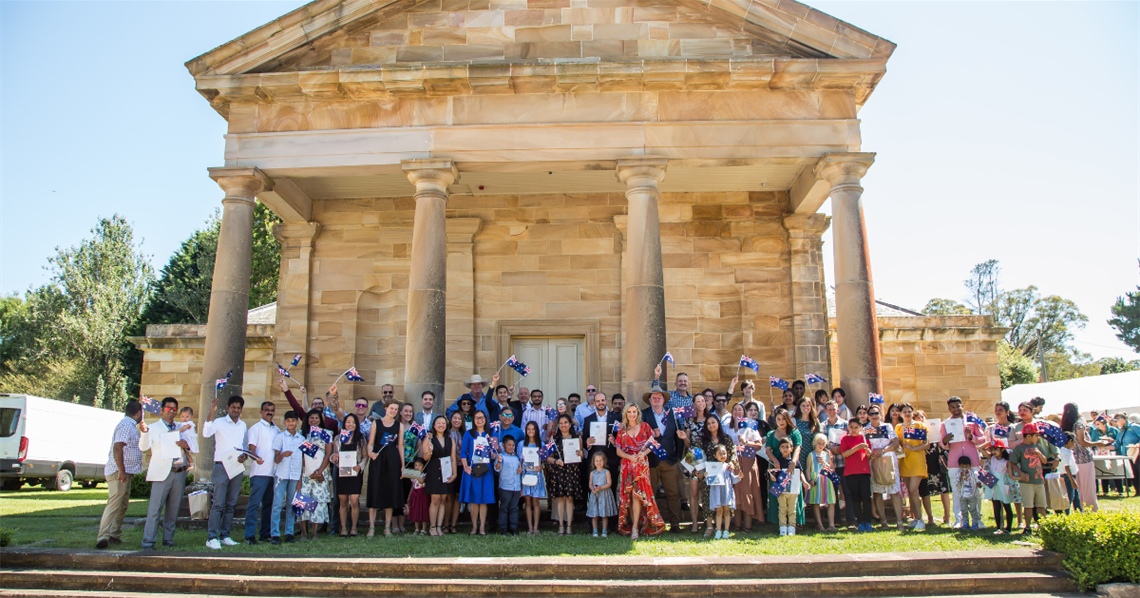 Large group of people standing in front of Berrima Court House waving Australian Flags