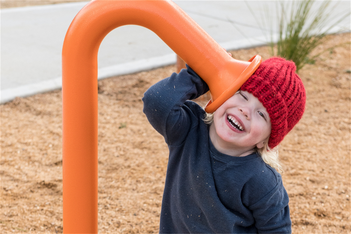 Young child in read beanie smiles listening with ear against play equipment