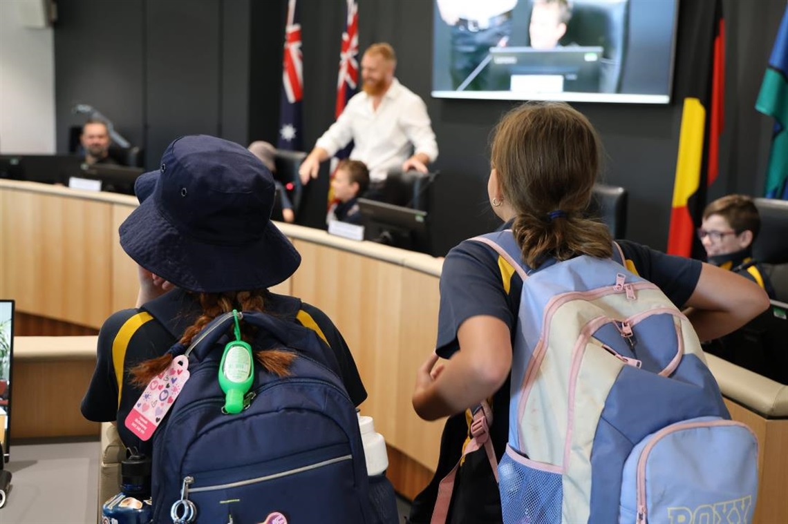 Two school students with backpacks watch presentation by Mayor Fitzpatrick in Council Chambers