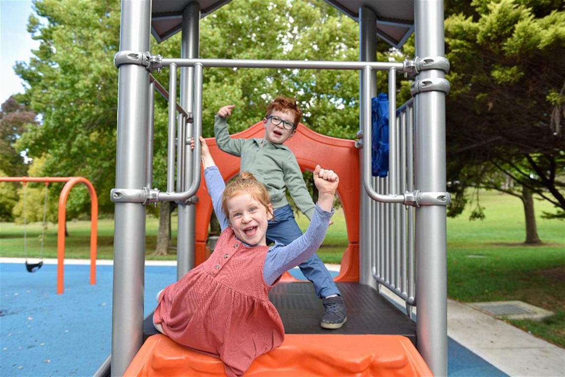 Photograph of two children on play equipment holding their arms up and smiling at camera 