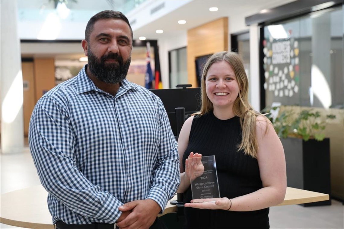 Photo of George and Rhianon from Council's Customer ExperienceTeam hold glass award smiling in front of customer service desk 