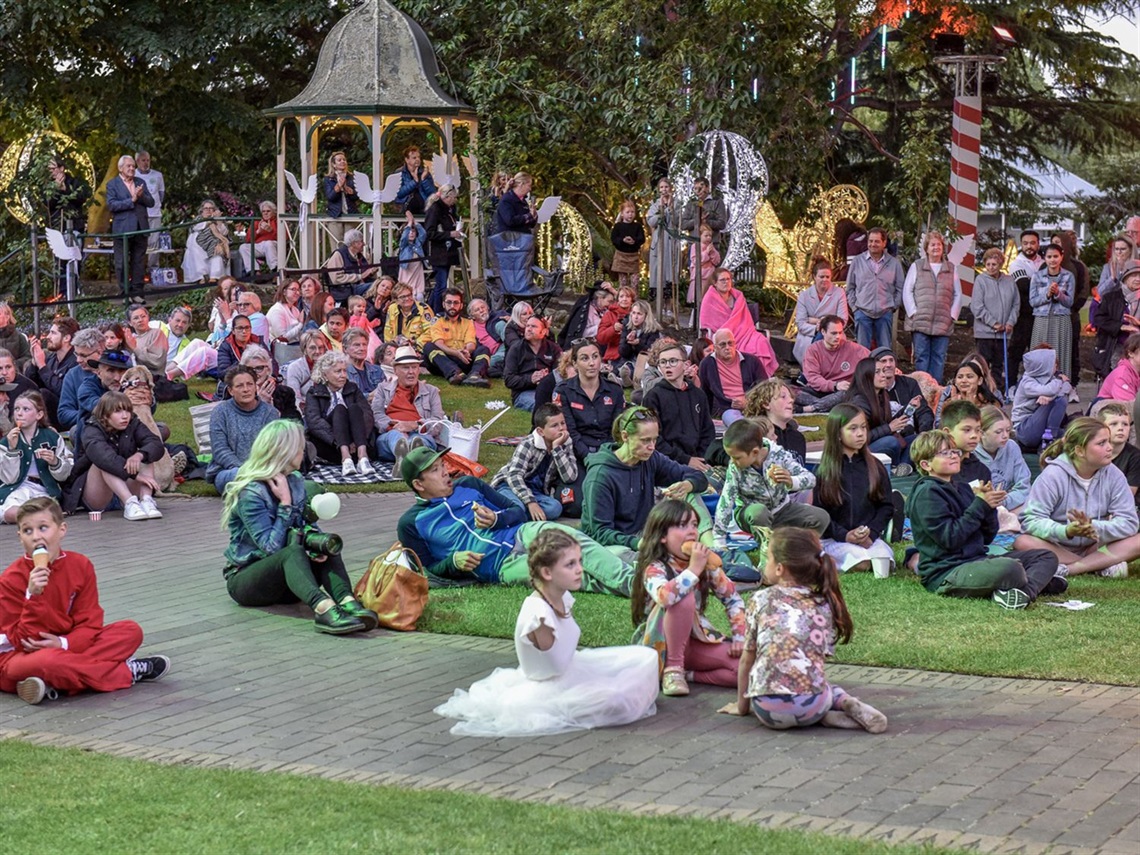 A large crowd of community members enjoy Christmas festivities in Corbett Gardens with Rotunda and festive angel wing decorations in background