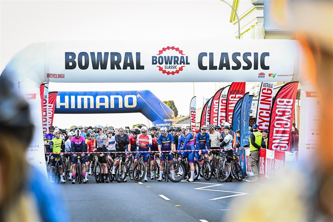 Bowral Classic Bike Riders on road starting line behind ribbon, road lined with red and white vertical banners