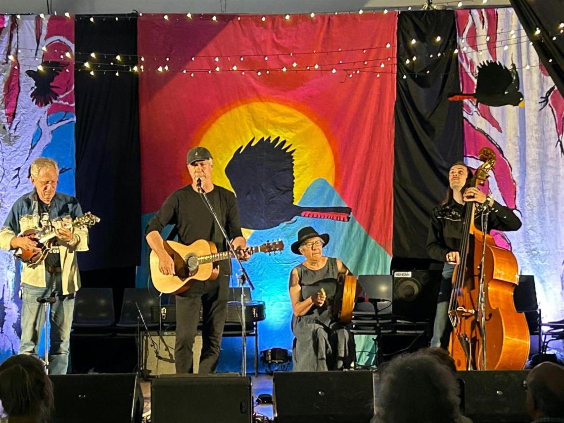 Four-piece band on stage in hall at Bundanoon Folk Festival 2023. Stage backdrop features colourful black cockatoo painting and black cockatoo themed decor