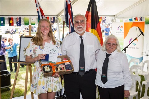 : Young Citizen of the Year 2024 - Mackenzie Isedale and Citizens of the Year 2024 - Uta Purcell and Dennis Purcell stand on stage in front of Australian, Aboriginal and Torres Strait Islander Flags with Australia Day Banner 