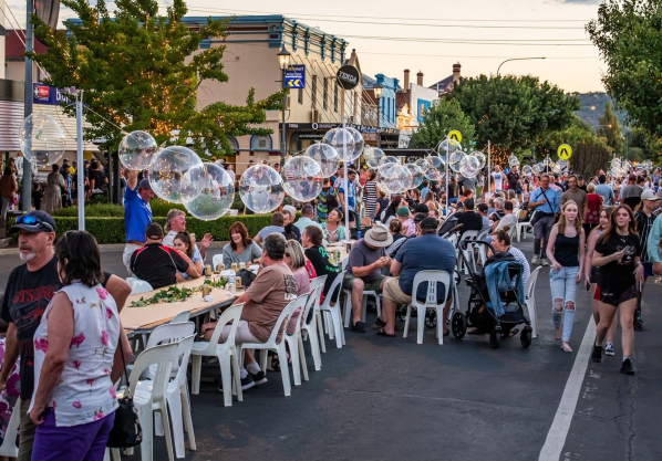 Community celebrating as part of outdoor Street Festival in town centre. An art installation hangs above long tables in middle of street like large bubbles. 