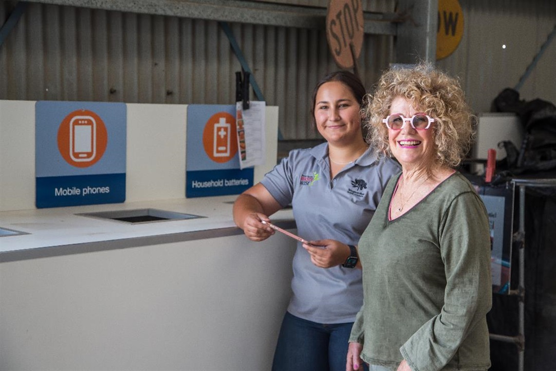 Two women, a customer and recycling centre staff member, stand at recycling drop off with mobile phone and battery signage in background