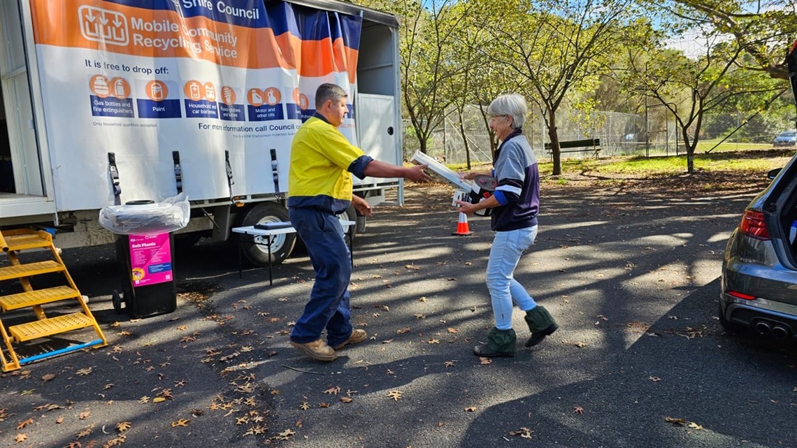 A staff member in high vis with Wingecaribee Shire Council's Mobile Community Recycling Service in background is passed paint tin, light tube and motor oil tin from a community member to be recycled