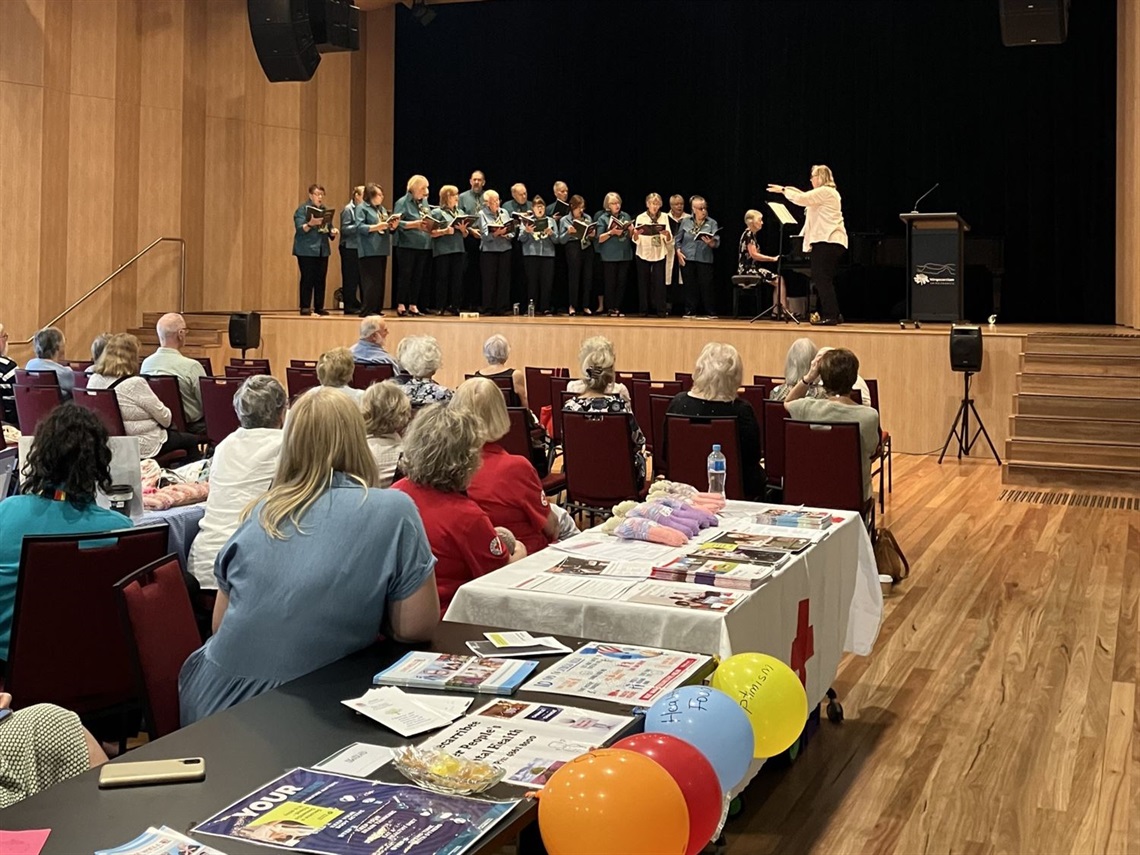 crowd in community hall with expo style tables watches choir on stage